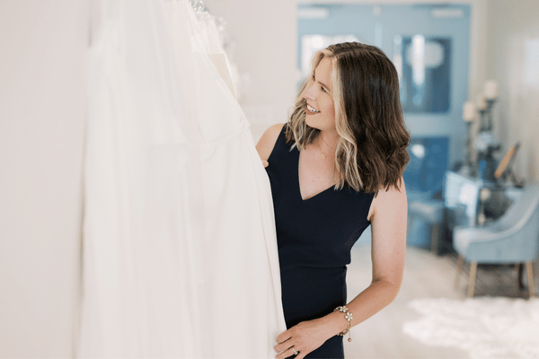 Smiling woman in a bridal boutique admires a flowing white wedding dress, conveying excitement and joy in a bright, elegant setting.