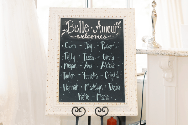 Framed chalkboard wedding sign reading &ldquo;Belle Amour welcomes,&rdquo; surrounded by guest names; elegant white decor creates a warm, romantic tone.