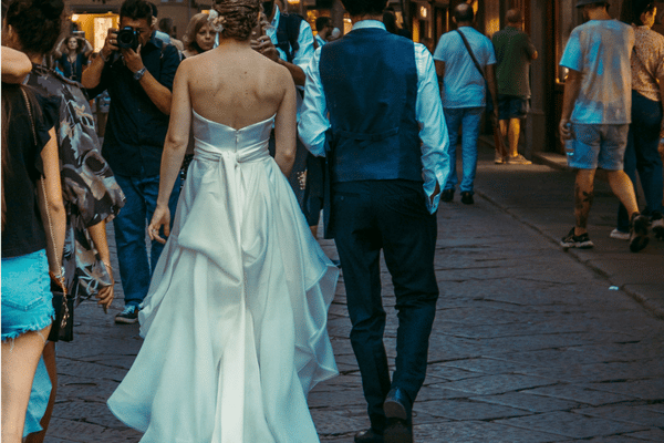 Bride in a strapless white gown and groom in a blue suit walk through a lively city street, surrounded by pedestrians in a candid wedding scene.
