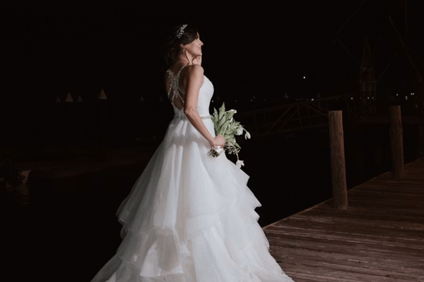 Bride in a flowing white wedding gown carries a bouquet along a wooden dock at night, creating an elegant, romantic, serene mood.