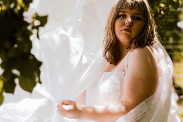 Portrait of a woman in a white dress holding sheer fabric outdoors, framed by leafy greenery; warm sunlight creates a serene, dreamy mood.