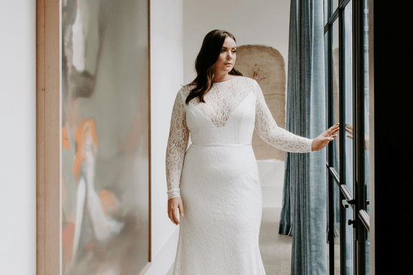 Bride in a long-sleeved white lace wedding gown stands by tall windows, touching the frame; soft light creates an elegant, serene mood.