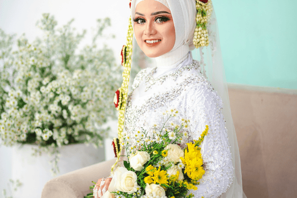 A smiling bride in a white dress and hijab, holding a bouquet of white and yellow flowers. 