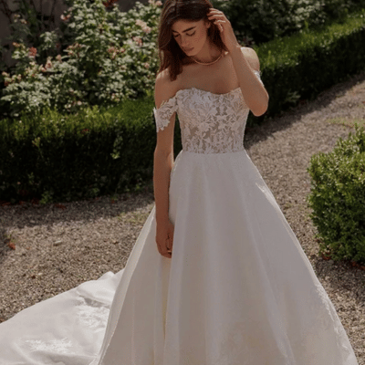 A woman wearing an elegant off-the-shoulder Aline wedding dress with floral lace details, standing in a garden. Explore more in our wedding dress style guide, discover bridal personality styles, and find unique wedding gowns to match your vision.