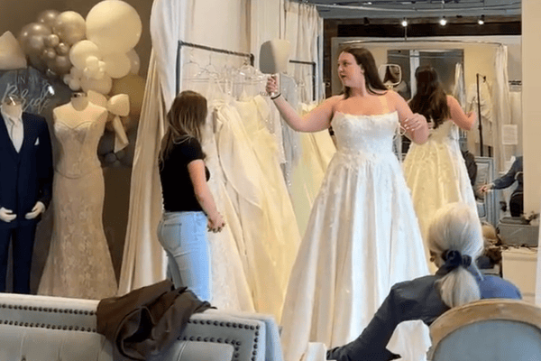 A bride-to-be trying on her white wedding dress gestures excitedly in a bridal shop. A consultant in jeans assists her while she checks the silhouette. The mood is joyful and anticipatory.