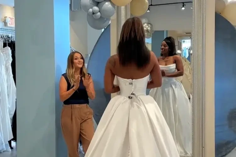 A bride in a strapless white wedding dress stands in front of a mirror, smiling. An excited wedding dress stylist in brown pants and a blue top claps nearby. Balloons decorate the background, creating a joyful, celebratory atmosphere.