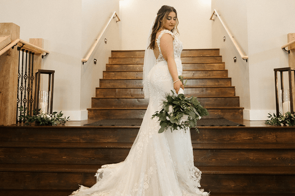 Bride in an elegant lace gown stands on wooden stairs, holding a bouquet thinking "no more wedding dress regrets". Soft lighting and floral decor create a romantic, serene atmosphere.