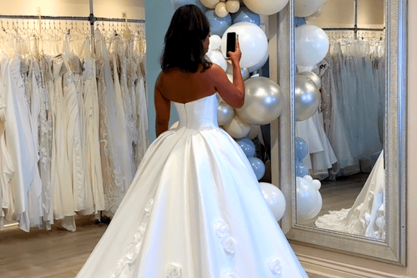 A woman in a bridal shop is trying on wedding dresses. She faces a mirror, her phone capturing the scene. Elegant ambiance.