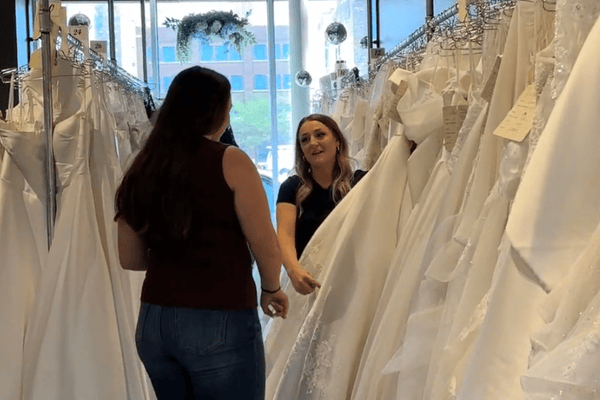 Two women in a bridal shop discuss wedding dresses. One holds a white gown, surrounded by racks of dresses, with a bright window behind them.