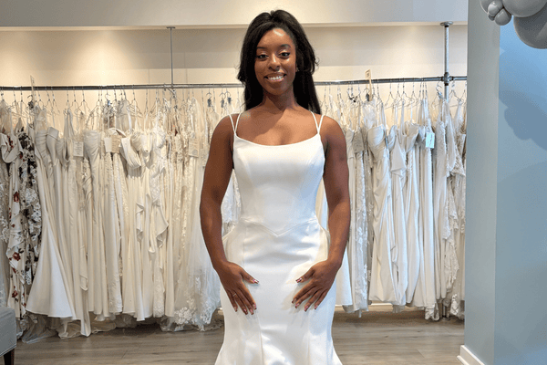 Bride standing in a bridal boutique wearing a sleek satin gown while exploring off the rack wedding dresses displayed behind her.
