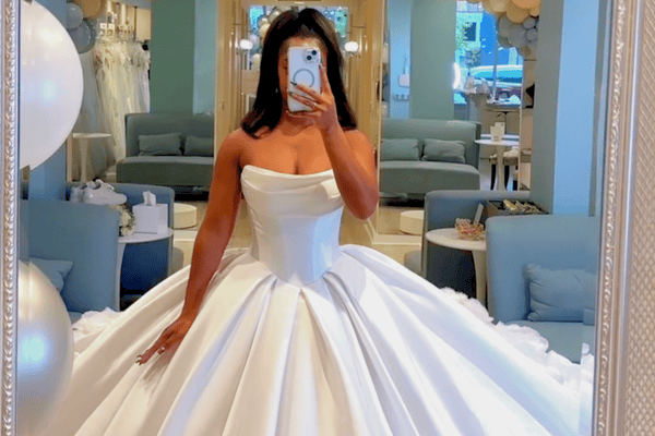 Woman in a bridal shop wearing a strapless, voluminous white wedding gown, taking a mirror selfie. The room is decorated elegantly with soft colors.
