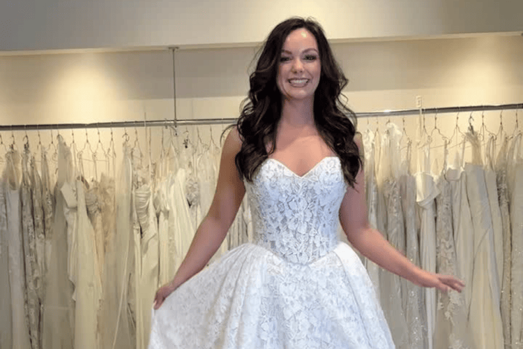 A smiling woman stands in a bridal shop wearing a white lace wedding gown. She holds the dress's skirt, surrounded by hanging dresses in soft lighting.