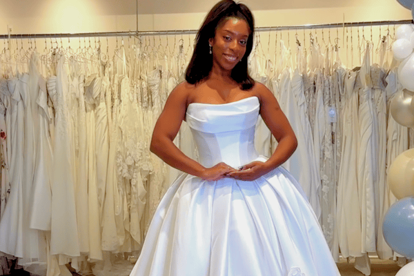 A woman in a bridal boutique wears a strapless, satin ball gown. She smiles confidently with hands clasped, surrounded by rows of white wedding dresses.
