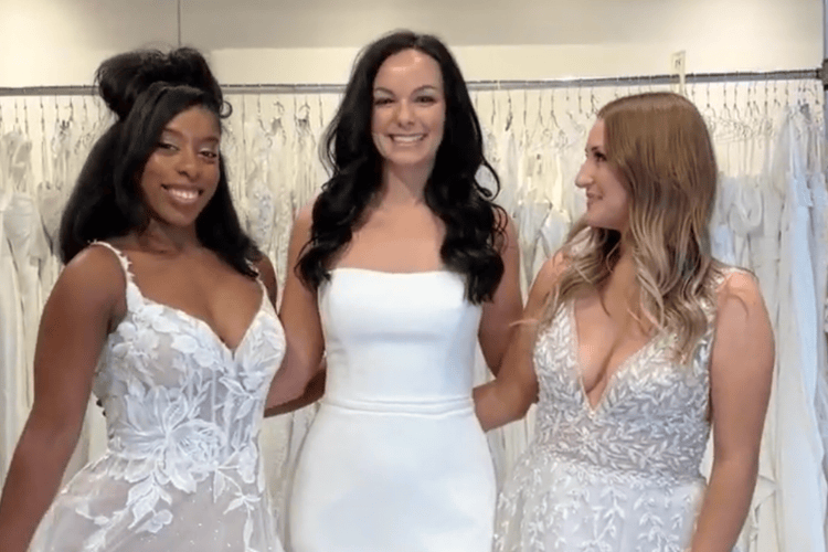 Three women in a bridal shop smile joyfully, each wearing a white wedding dress with intricate lace designs, surrounded by hanging gowns.