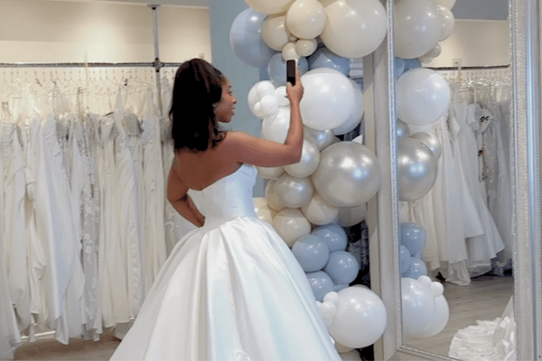 A woman in a strapless white wedding gown captures a selfie in front of a mirror adorned with white and silver balloons, surrounded by wedding dresses.
