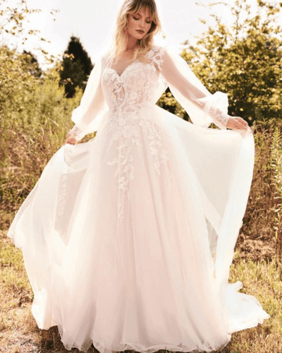 A bride in a flowing white wedding gown with lace details, standing in a grassy field. 