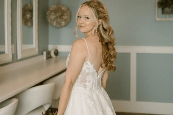 A bride with wavy blonde hair smiles over her shoulder in an elegant white dress with lace details. The room is softly lit, conveying a serene mood.