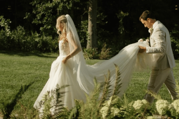 Bride and groom walking on grass; the groom holds the bride's long, flowing veil. Sunlight filters through surrounding trees, creating a serene ambiance.