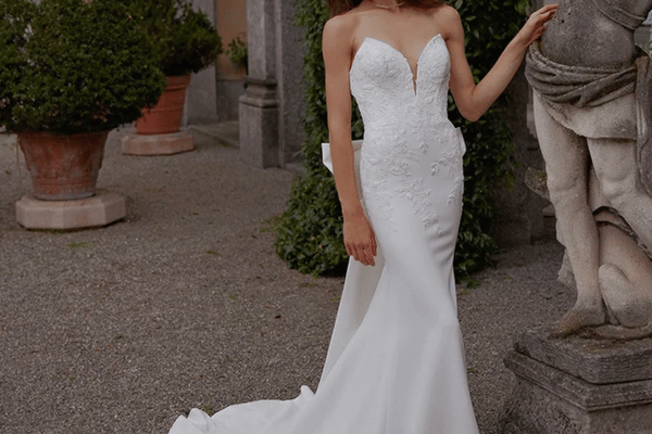Petite bride wearing a strapless fit-and-flare wedding dress with floral lace detailing and a sweetheart neckline, standing outdoors beside a stone statue.