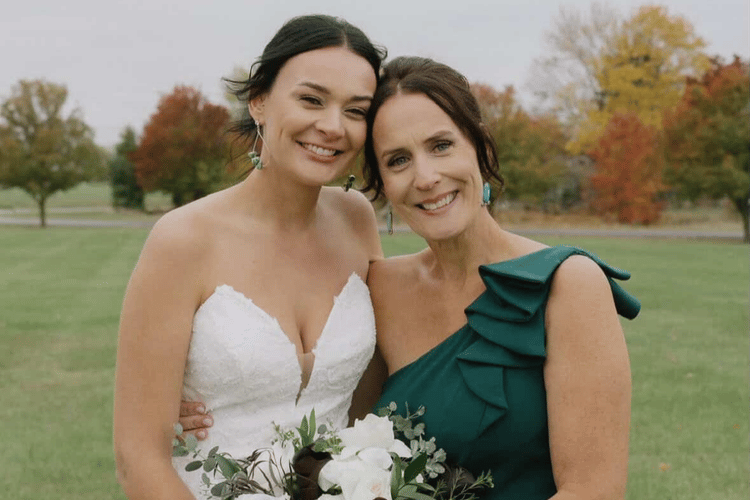 Mother of the bride posing with the bride outdoors in an elegant emerald green one-shoulder dress during a fall wedding celebration.