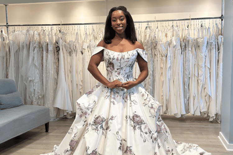 A bride smiles in a bridal shop, wearing an off-shoulder wedding gown with floral patterns. She stands confidently among rows of hanging dresses, smiling at the wedding dress stylist.