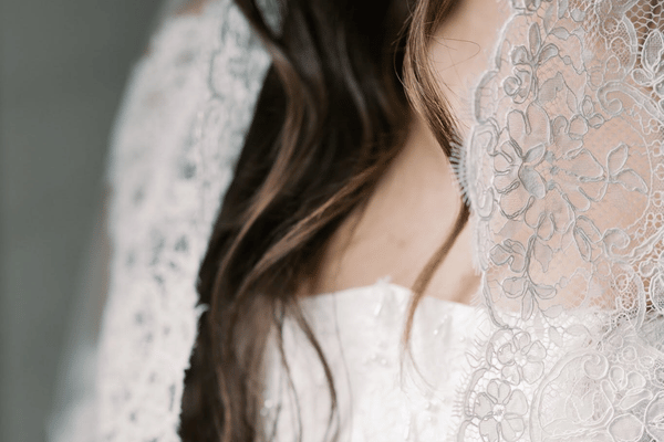 Close-up detail of a bride wearing a lace wedding veil over a white wedding dress, highlighting delicate floral lace patterns and soft curls.