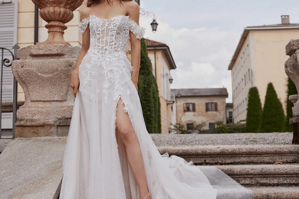 Bride wearing an off-the-shoulder A-line wedding dress with floral lace appliqu&eacute;s, a flowing skirt, and a thigh-high slit, photographed on stone steps.