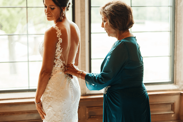 Mother of the bride helping fasten the bride&rsquo;s wedding dress in a softly lit room