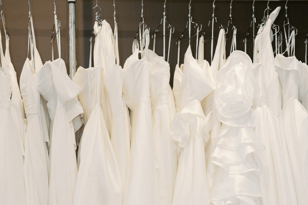 A collection of white wedding dresses hanging on racks in a bridal boutique, featuring a variety of silhouettes, necklines, and fabric details.