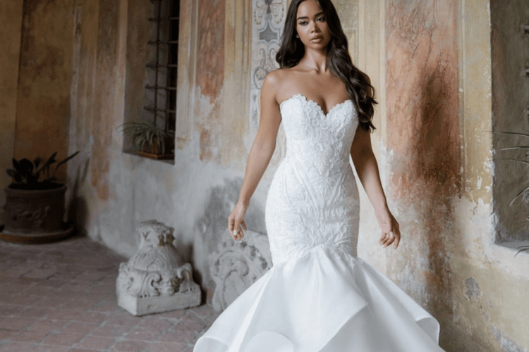 A bride in a strapless, lace-embroidered mermaid wedding dress stands confidently in a rustic hallway with textured walls and potted plants.