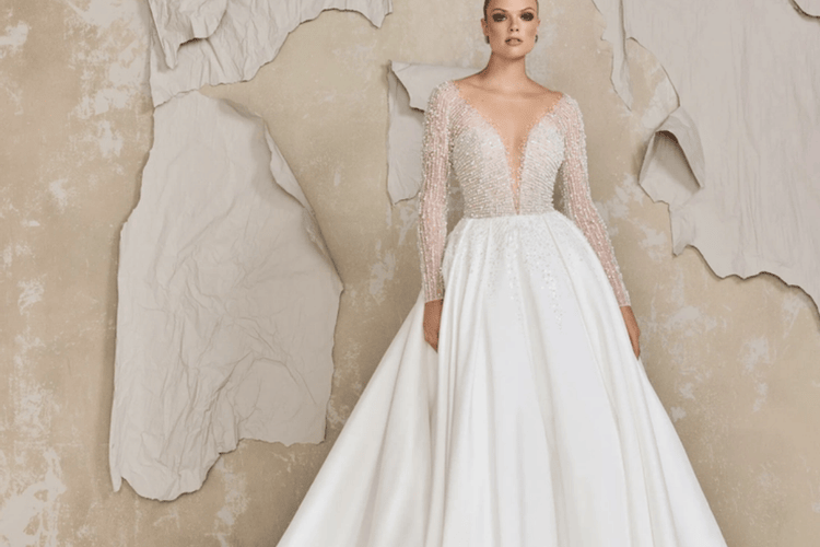 A woman in an elegant wedding gown with a sheer, beaded top and full skirt stands confidently. The textured wall backdrop adds a modern contrast.
