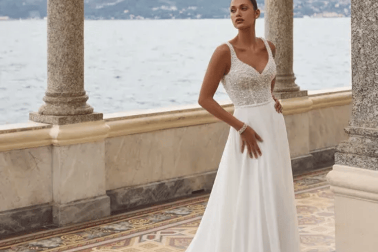 A woman in an elegant white wedding dress with a beaded bodice stands on a tiled balcony overlooking a serene body of water, evoking a romantic and serene tone.