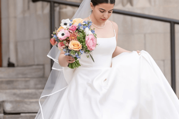 A bride in a minimalist wedding dress walks down steps, holding a colorful bouquet of flowers.