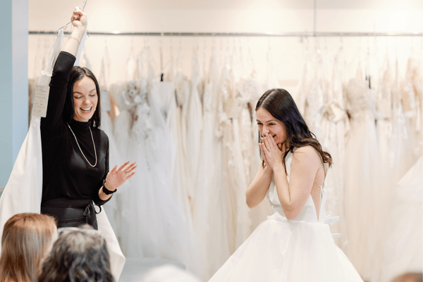 A smiling bride in a white dress covers her mouth as a stylist raises a dress tag, surrounded by bridal gowns in the background—a perfect reminder of what to bring wedding dress shopping for a stress-free and joyful experience.