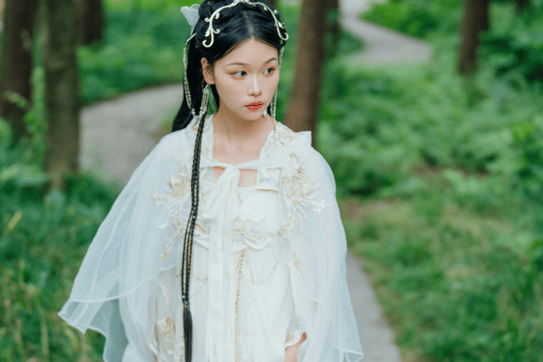 A young woman in a white dress and flowing cape, walking on a path in a lush green forest. 