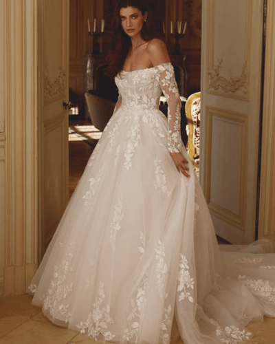 A model in a stunning off-shoulder wedding dress with floral lace designs, standing in an elegant room. 