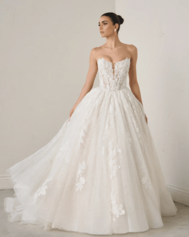 A model wearing a strapless white wedding gown with floral details, standing gracefully. 