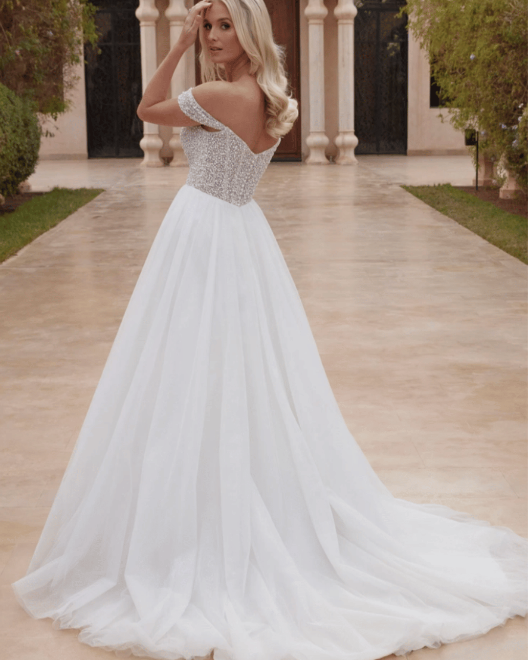 A woman in an off-the-shoulder, white wedding dress with a long train, posing outdoors.