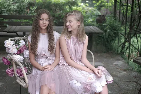 Two girls in blush pink dresses sitting together on a wicker chair beside a floral arrangement in a garden setting.