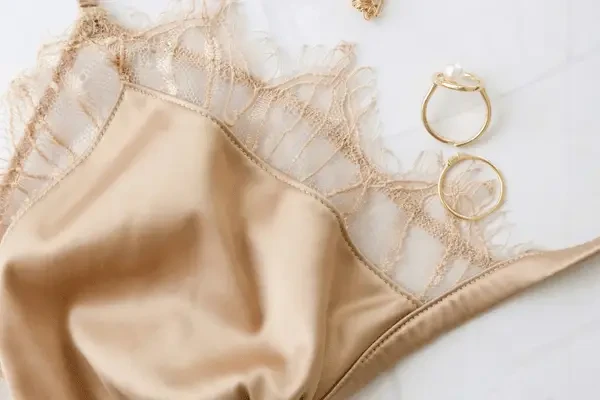 Champagne-colored satin and lace bra with delicate trim displayed on a white surface beside two gold rings.