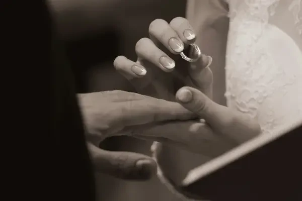 Close-up black-and-white photograph of a bride receiving a wedding ring on her finger, with her lace wedding dress visible in the background.