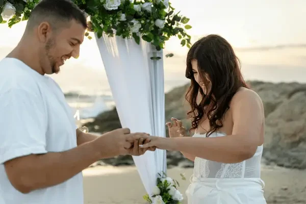 Bride and groom exchanging rings during a beach wedding ceremony beneath a floral arch, with the ocean and sunset in the background.