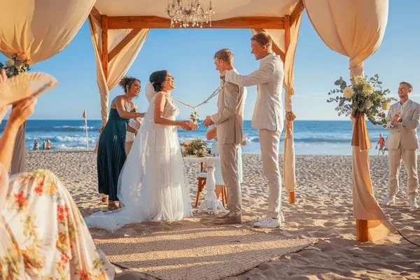 Bride and groom standing beneath a decorated beach wedding arch during their ceremony, surrounded by family and friends with the ocean in the background.