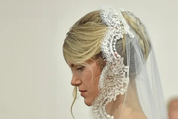 A bride in profile wearing a lace-trimmed wedding veil over a blonde hairstyle, with delicate lace detailing framing her face.