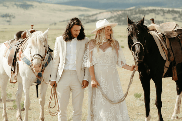 A bride in a white dress and a groom in a light suit hold hands by two horses in a field. 