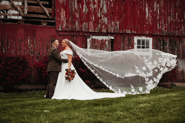 A bride and groom share a moment outside a rustic red barn, the bride's veil, a stunning bridal accessory, flowing behind her.