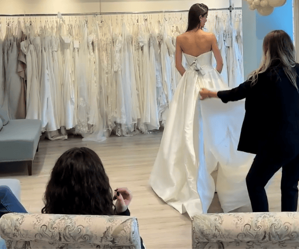 A bride in a white gown with a bow is being adjusted by a stylist, while two women watch from a couch. 