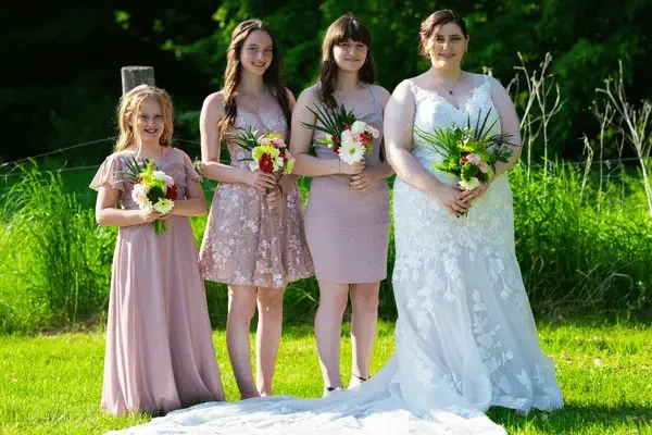 Young flower girl in a pink dress carrying a basket of flowers while walking along an outdoor wooden bridge.