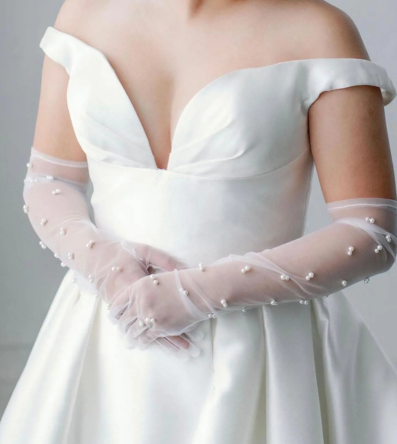 A close-up of a bride wearing a white satin wedding dress with an off-the-shoulder neckline and sheer pearl-embellished gloves. The elegant gown highlights the importance of proper care and storage to maintain its beauty after the wedding day.