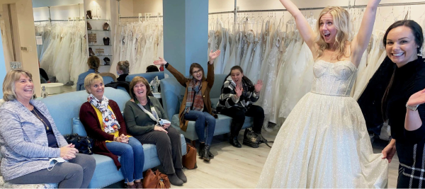 A joyful bride-to-be celebrates while trying on a wedding dress, surrounded by her supportive family and friends in a bridal boutique. Discover who to take with you wedding dress shopping for a fun and memorable experience.